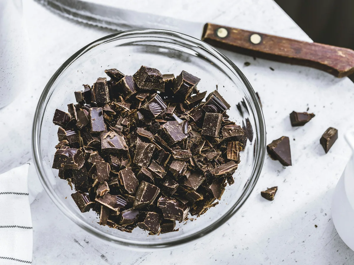 Chopped dark chocolate fills a glass bowl beside a kitchen knife.