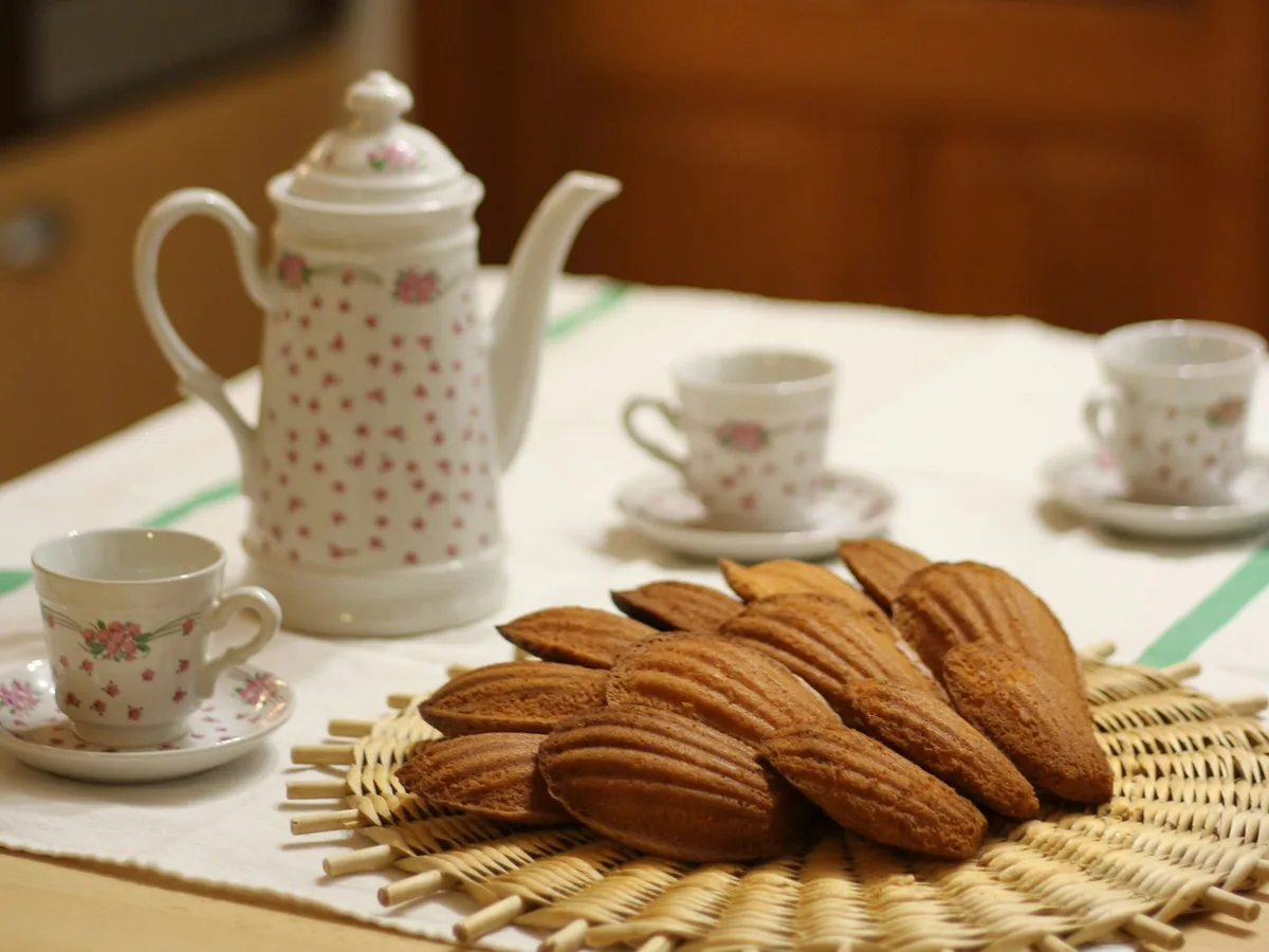A basket of madeleines sits beside a floral teapot and teacups.