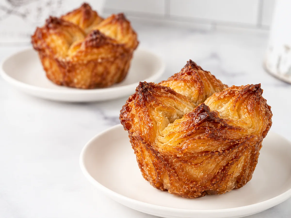 Two individual caramelized kouign-amann with four folded corners and crisp amber edges on white plates.